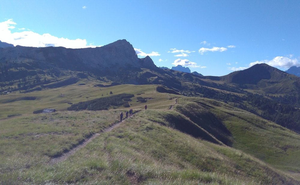 Gente caminando por un sendero entre las montañas, en un día soleado. Esto simboliza el camino de la vida, por el que a veces tenemos que superar obstáculos tan grandes como montañas, pero siempre avanzando hacia la luz del sol.